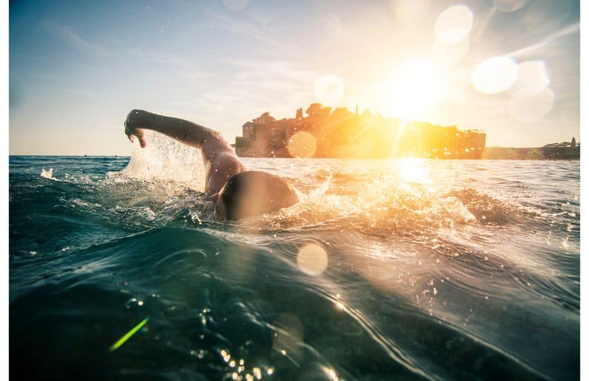 Man swimming with the blazing sun behind him as he swims away from shore.